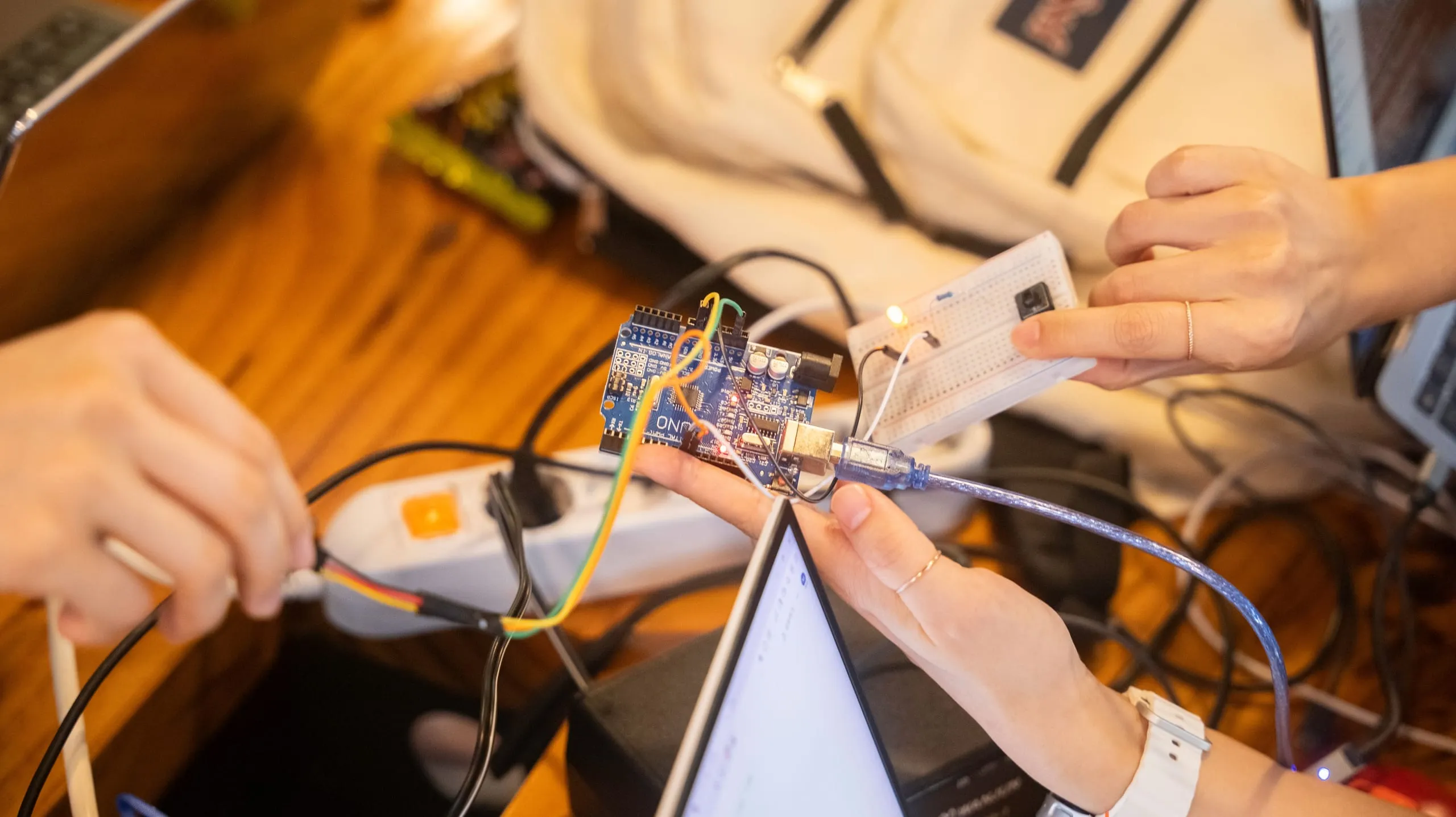Due persone lavorano insieme a un tavolo: una tiene in mano una scheda Arduino collegata con il cavo USB al portatile, l'altra una breadboard con un LED acceso