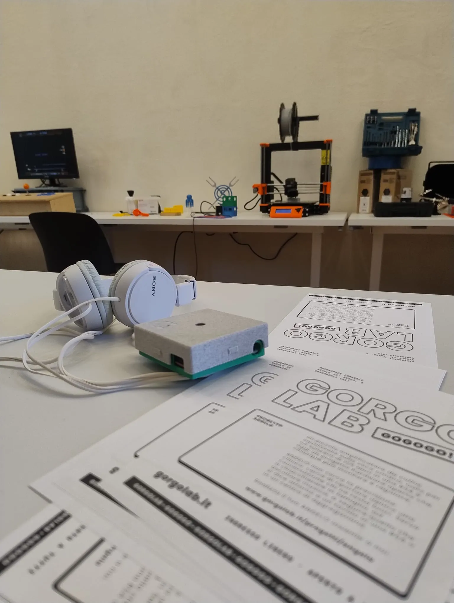 Un paio di cuffie bianche e un piccolo contenitore stampato in 3D, grigio e verde, appoggiati su un tavolo accanto ai volantini del Gorgo Lab; sullo sfondo una stampante 3D e altri progetti