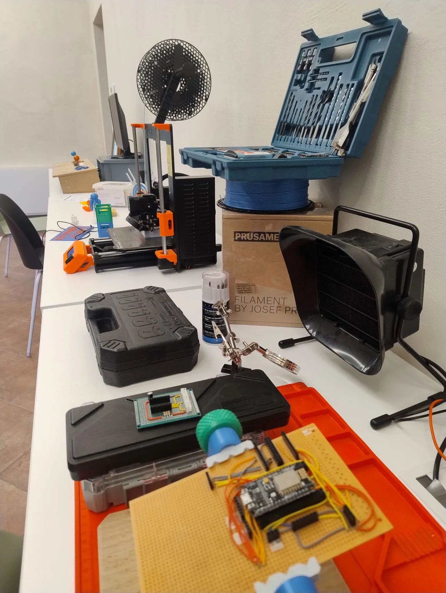 Un banco di lavoro con un circuito millefori e una scheda a microcontrollore in primo piano, una terza mano con lente, un aspiratore per i fumi di saldatura, valigette di attrezzi e la stampante 3D sullo sfondo