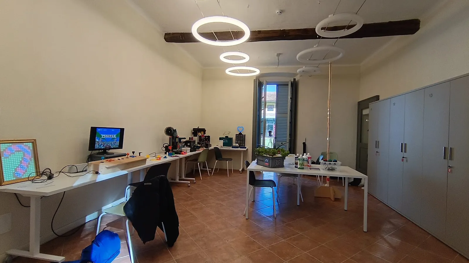 La sala del Gorgo Lab a Ca' Busca, con lampade ad anello sotto le travi di legno, lunghi tavoli bianchi carichi di progetti e attrezzi e una cassetta di piante sul tavolo centrale