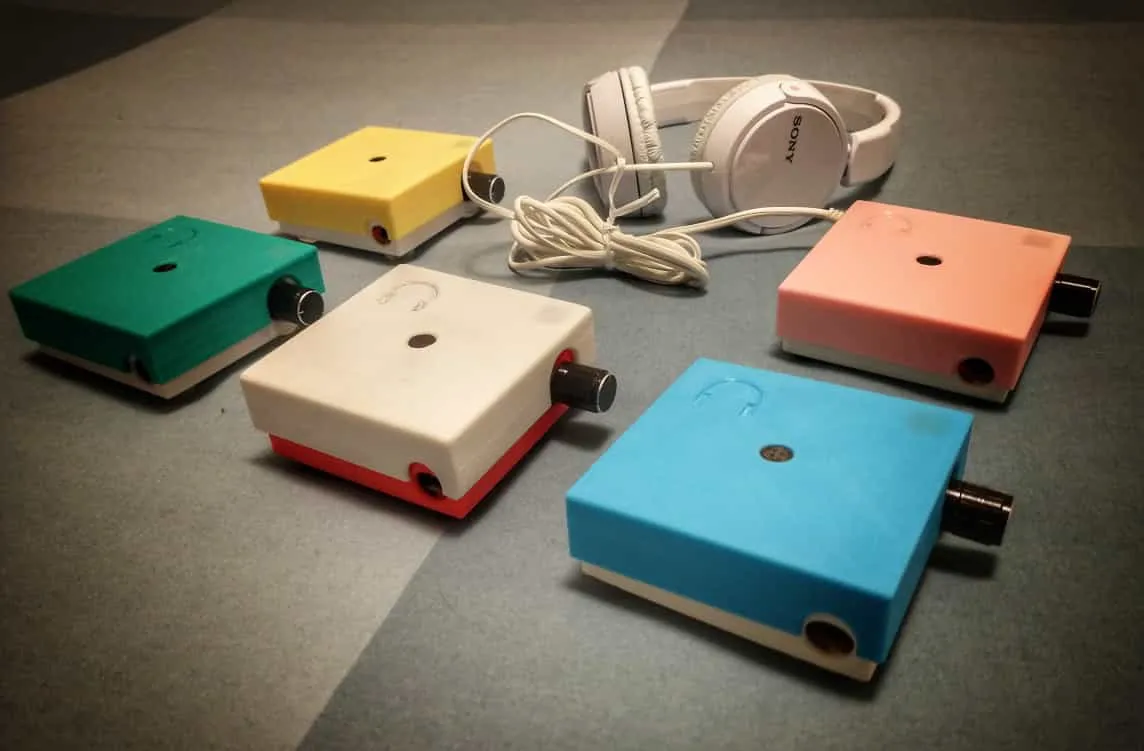 Cinque ANGELO con il contenitore stampato in 3D in colori diversi, giallo, verde, bianco, rosa e azzurro, appoggiati su un tavolo accanto a un paio di cuffie bianche