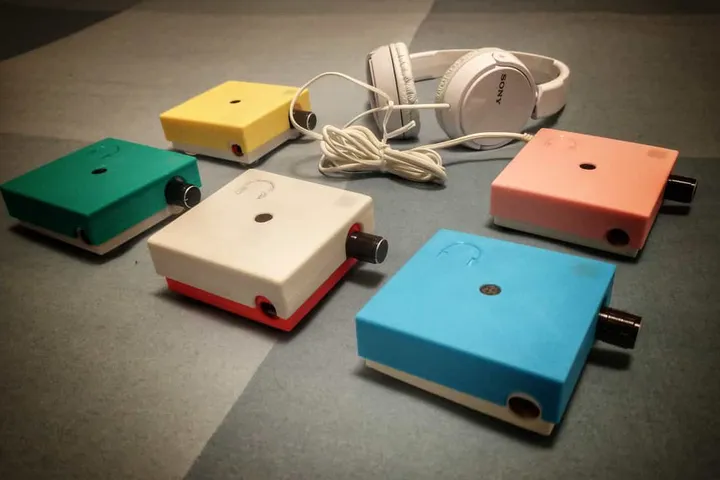 Cinque ANGELO con il contenitore stampato in 3D in colori diversi, giallo, verde, bianco, rosa e azzurro, appoggiati su un tavolo accanto a un paio di cuffie bianche