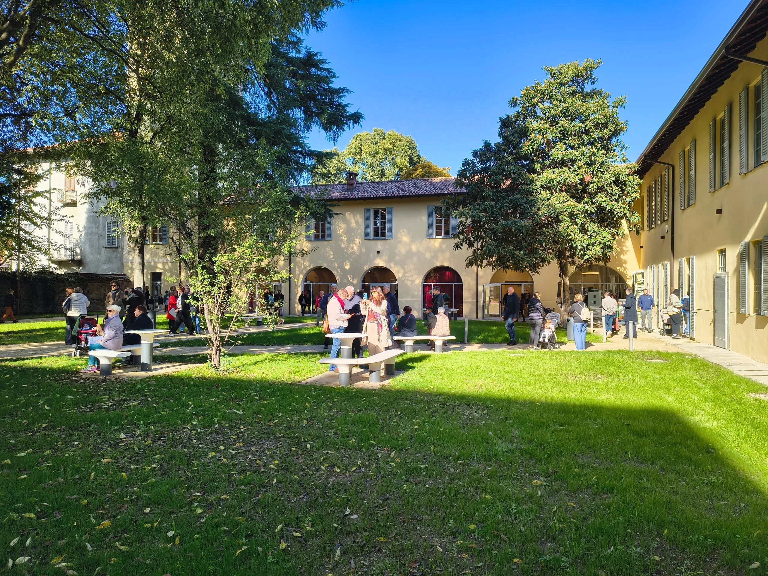 Il cortile di Ca' Busca in una giornata di sole: un prato verde con alberi alti, panchine e tavolini bianchi, gruppi di persone che passeggiano davanti all'edificio giallo con i portici ad arco