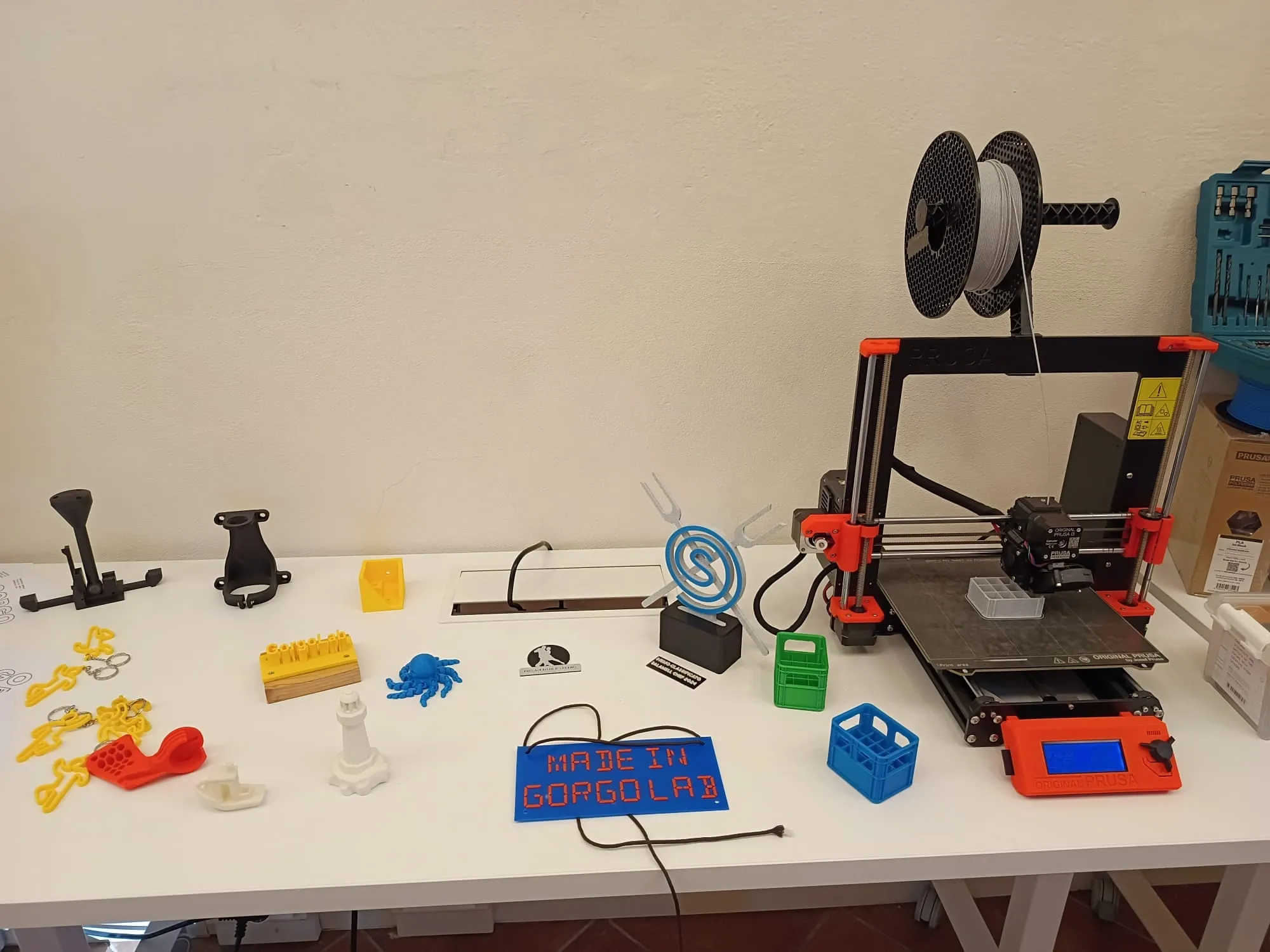 Una stampante 3D arancione e nera su un tavolo, circondata da oggetti stampati: portachiavi gialli, un polpo azzurro, una barchetta bianca, un faro, cassette in miniatura e una targa blu con la scritta "Made in Gorgolab"